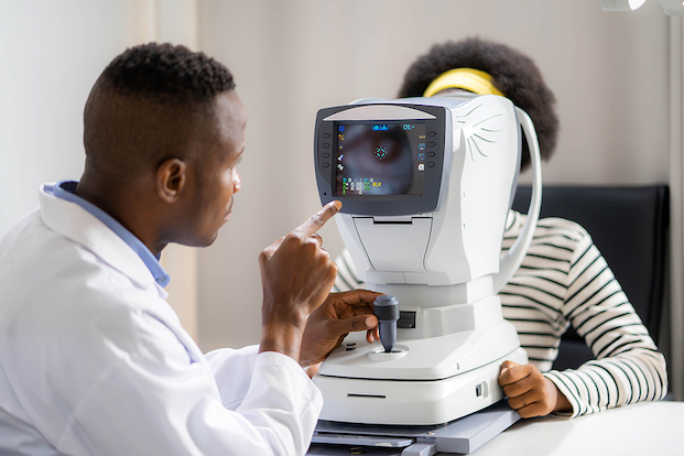 Brown skinned man or doctor interpreting results on a vision machine, while brown skinned woman looks into the machine. 